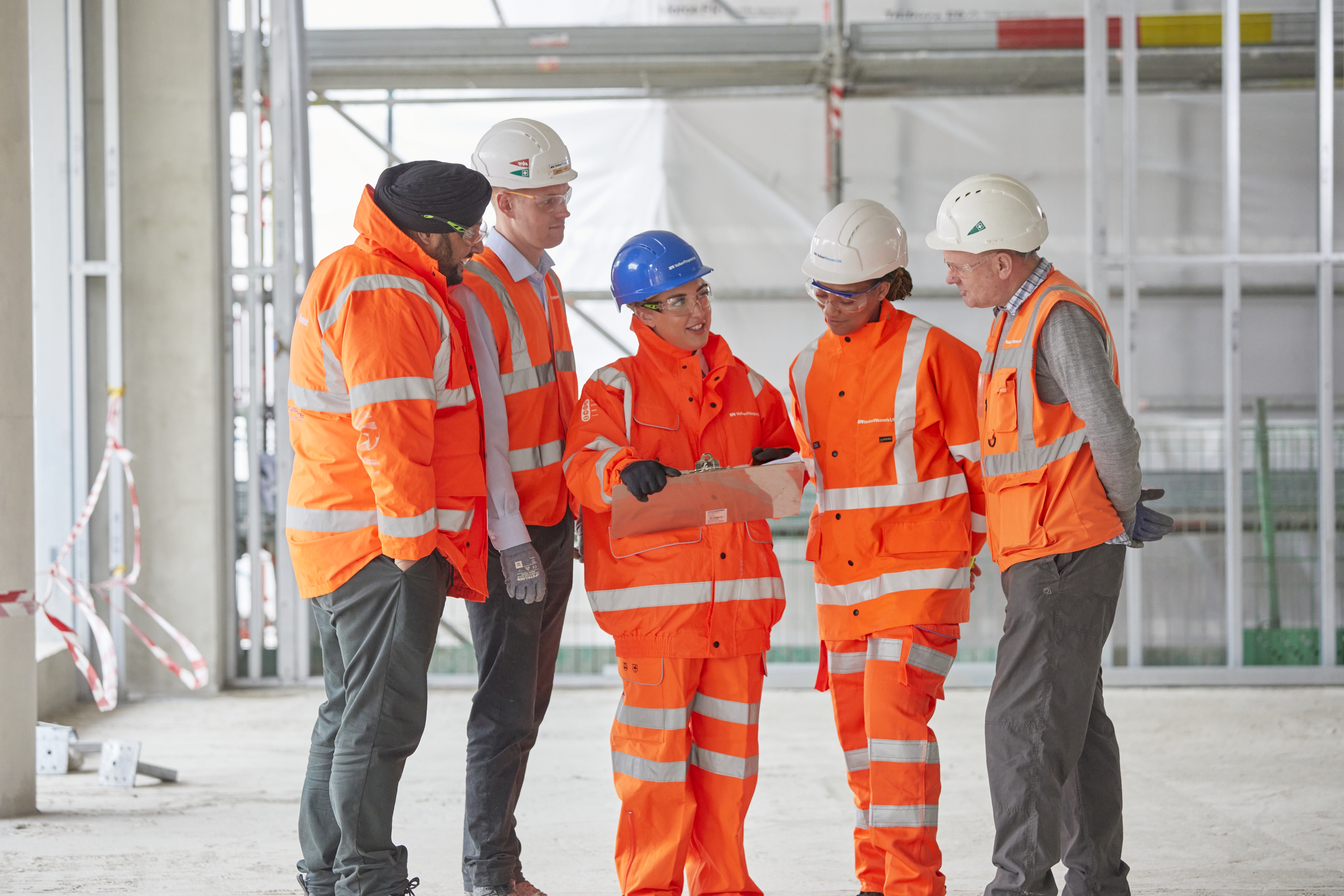  A diverse mixture of women and men in orange high-vis clothing are on a site discussing documents in daylight.